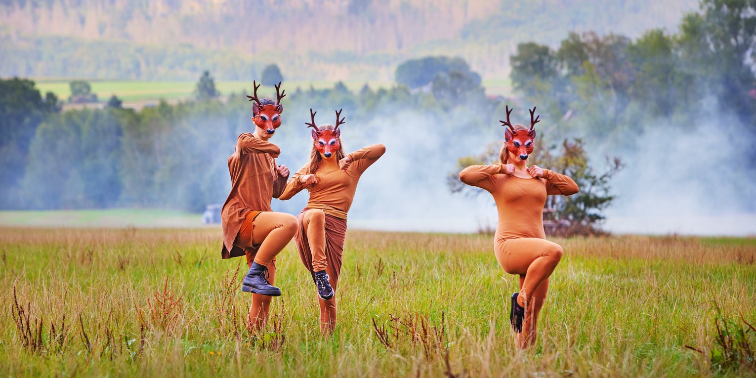 Drei Menschen in Rehkostümen tanzen in einem Feld, im Hintergrund ein Wald und Nebelschwaden.