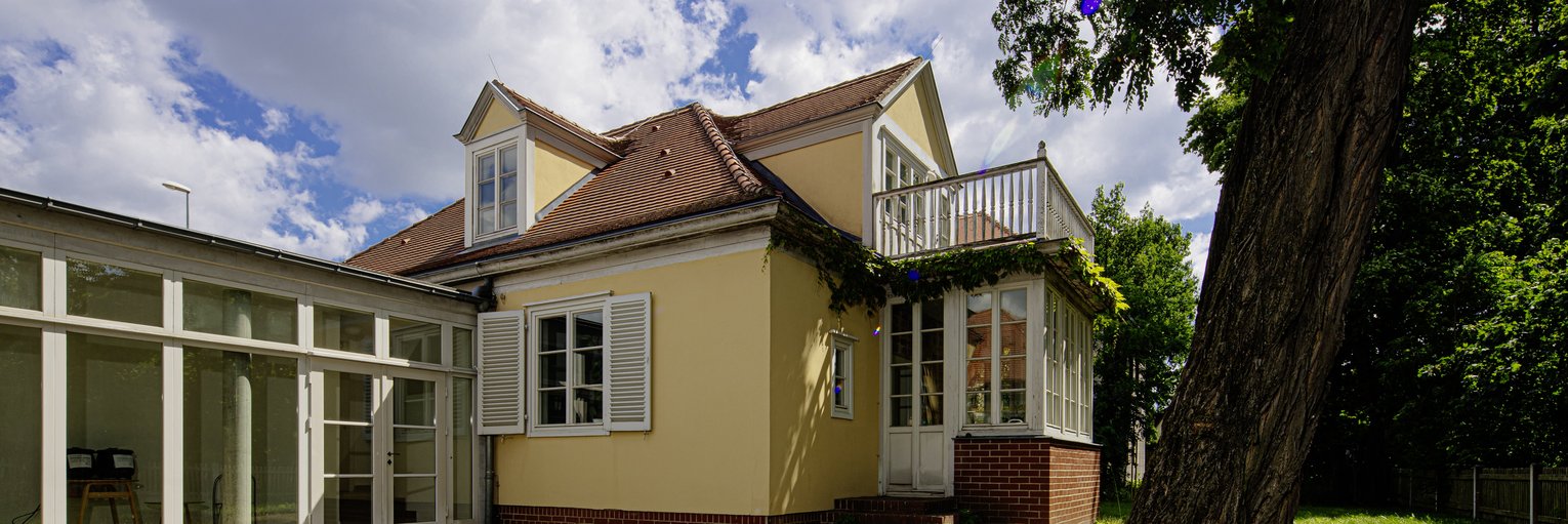 Ein gelbes Haus mit einer Dachgaube, weißen Fensterläden und einem gläsernen Wintergarten, im Vordergrund eine Wiese mit Baum.