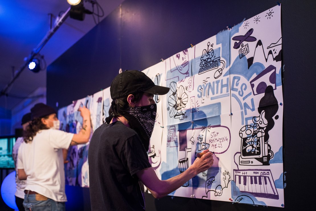 An einer schwarzen Wand ist weißes Papier mit Pins befestigt. Darauf schreiben Jugendliche mit schwarzen und blauen Markern Schriftzüge und zeichnen Comicfiguren.