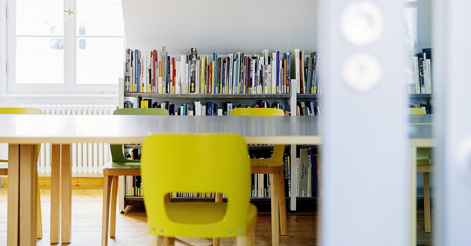 Vor einem an der Wand verlaufenden Bücherregal steht ein langer Tisch mit grünen Stühlen. 