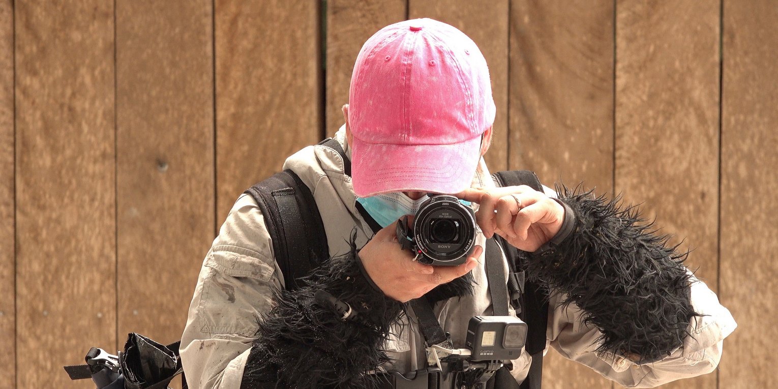 Ein Mann mit pinkem Basecap und braunem Fellanzug hält frontal eine Kamera. 