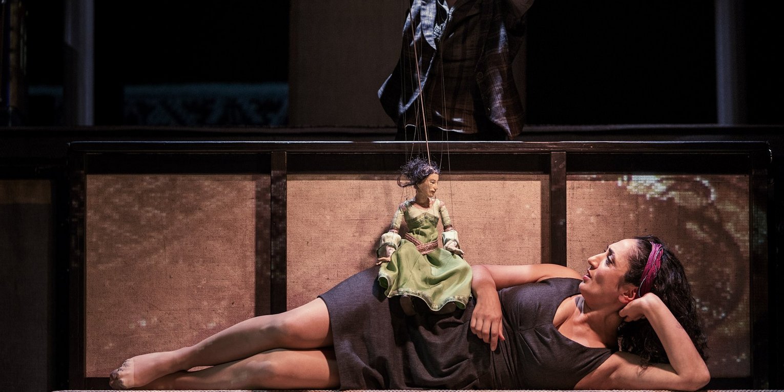 Eine Schauspielerin liegt seitlings auf dem Boden. Auf ihrer Hüfte sitzt eine grün und festlich gekleidete Marionettenpuppe, die von einer dahinterstehenden Frau gespielt wird.